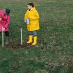 Dois voluntários plantam árvores em um campo gramado, usando capas de chuva coloridas e botas.
