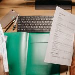 Mãos analisam planilhas financeiras sobre uma mesa, ao lado de um teclado e materiais de escritório.