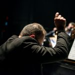 Maestro conduz uma orquestra sinfônica durante apresentação em uma sala de concertos, com músicos e partituras visíveis.