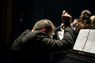 Maestro conduz uma orquestra sinfônica durante apresentação em uma sala de concertos, com músicos e partituras visíveis.