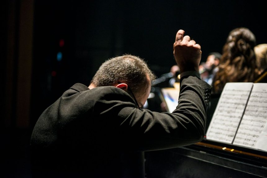 Maestro conduz uma orquestra sinfônica durante apresentação em uma sala de concertos, com músicos e partituras visíveis.