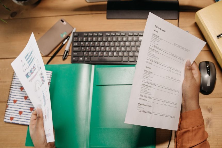 Mãos analisam planilhas financeiras sobre uma mesa, ao lado de um teclado e materiais de escritório.